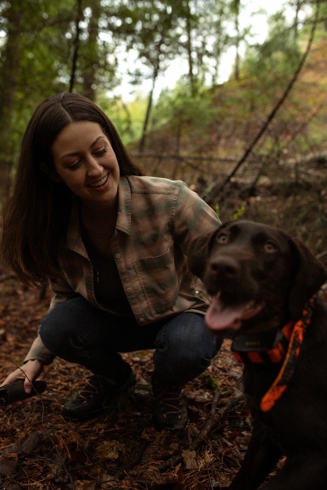 Tara Vancil is crouching down in the woods smiling as she looks affectionately at her dog Maven.